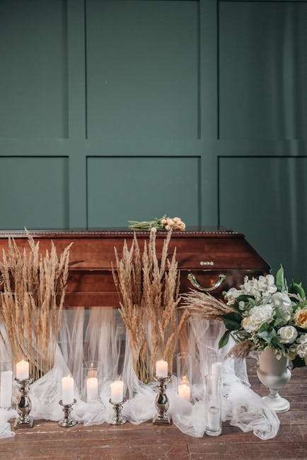 A beautifully arranged floral tribute featuring white roses, lilies, and soft pastel carnations in a white ceramic vase, accompanied by tall, dried pampas grass accents. The bouquet is positioned on a