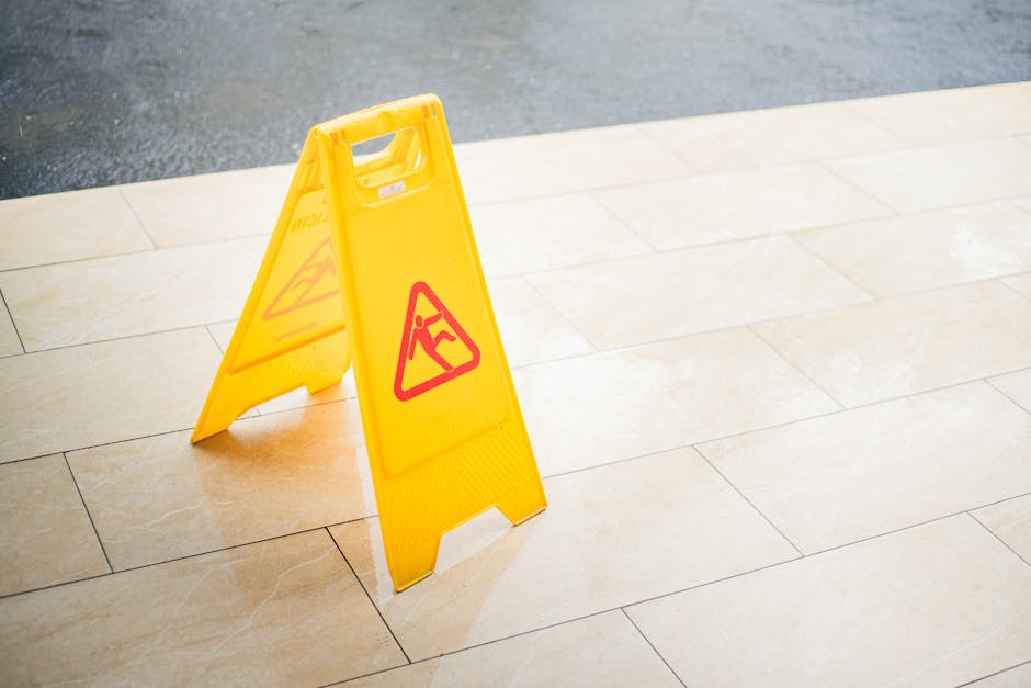 A yellow 'wet floor' caution sign with red and black triangular symbols stands on a beige tiled floor, indicating a potential slipping hazard. The sign is positioned near the edge of the tiled area, w