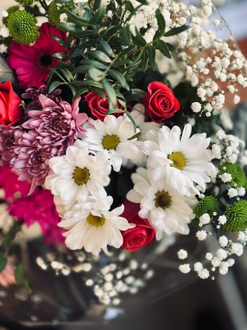 A close-up of a floral bouquet featuring white daisies with bright yellow centers, deep pink roses, and purple chrysanthemums, accented with delicate sprigs of white baby's breath and glossy green fol