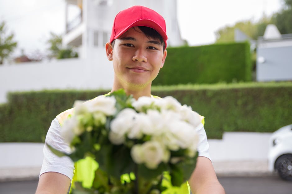 A young male flower delivery courier with light skin, wearing a red cap and a reflective high-visibility vest, is standing outdoors on a residential street during daytime, smiling gently as he extends
