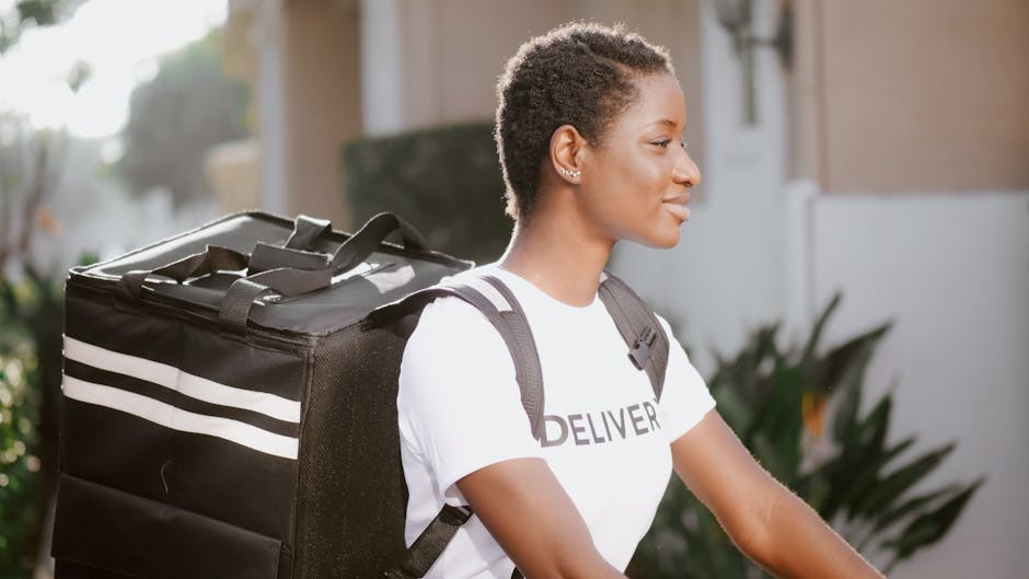 A young woman with short, textured hair is outdoors, wearing a white T-shirt with the word 'DELIVERY' partially visible, and carrying a large black insulated delivery bag with white stripes over her s