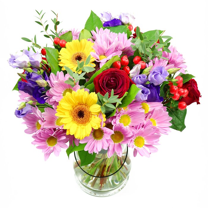 A lush bouquet of mixed blooms stands proudly in a simple clear glass vase, framed by a pure white backdrop that lets each colour and texture shine. At the core, velvety red roses curve open in layered spirals, offering an immediate sense of romance and depth, like a classic gesture of affection delivered to a doorstep in Ardleigh Green. Near them, large yellow gerbera daisies with deep, warm centres beam outward, their sunny petals acting as bright focal points that infuse the arrangement with cheerful energy. Surrounding this central grouping, clusters of pink and pale lavender chrysanthemums or daisies spill generously outward, their many fine petals and tiny yellow centres adding soft volume and a soothing, almost cloud-like quality. Threaded delicately among them are lisianthus blooms in shades of purple and lilac, their bell-shaped flowers and gently ruffled edges creating an elegant, cool-toned contrast that feels sophisticated yet approachable. Tucked between petals and foliage, small round hypericum berries in a vivid red bring playful texture and a modern twist, like little jewels hidden within the bouquet. The design is wrapped in layers of greenery, with broad, dark green leaves providing structure and depth, and lighter, more slender stems adding lift and movement, as if the flowers were caught in a gentle breeze over nearby Emerson Park. Through the clear vase, the neatly trimmed stems and fresh water are clearly visible, underscoring the care and precision of a skilled local florist. The composition speaks of joy, thoughtfulness, and celebration, perfect as a birthday surprise, a thank-you gesture around Ardleigh Green, or a welcoming centrepiece on a kitchen table bathed in afternoon light.